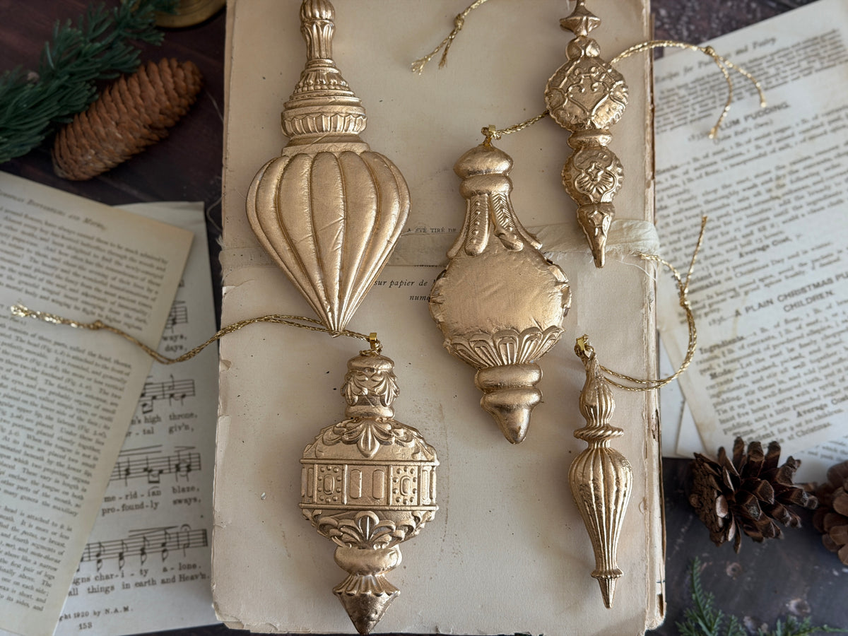 Five gold ornaments from the I.O.D. Baubles Mould (2026 Limited Release) displayed on a book with pinecones nearby.
