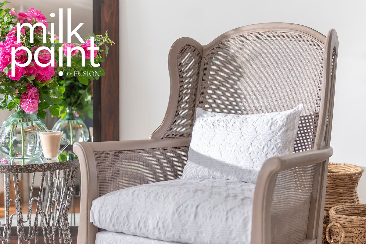 Wingback chair with white cushions next to round table; āAlmond Latte Milk Paint by Fusionā text above.