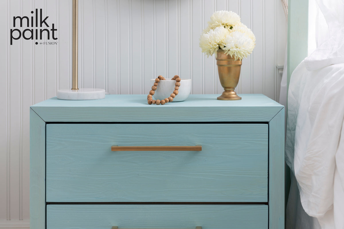 Nightstand in Fusion Amalfi Coast Milk Paint with gold handle, beaded bracelet, lamp, and white flowers on top.