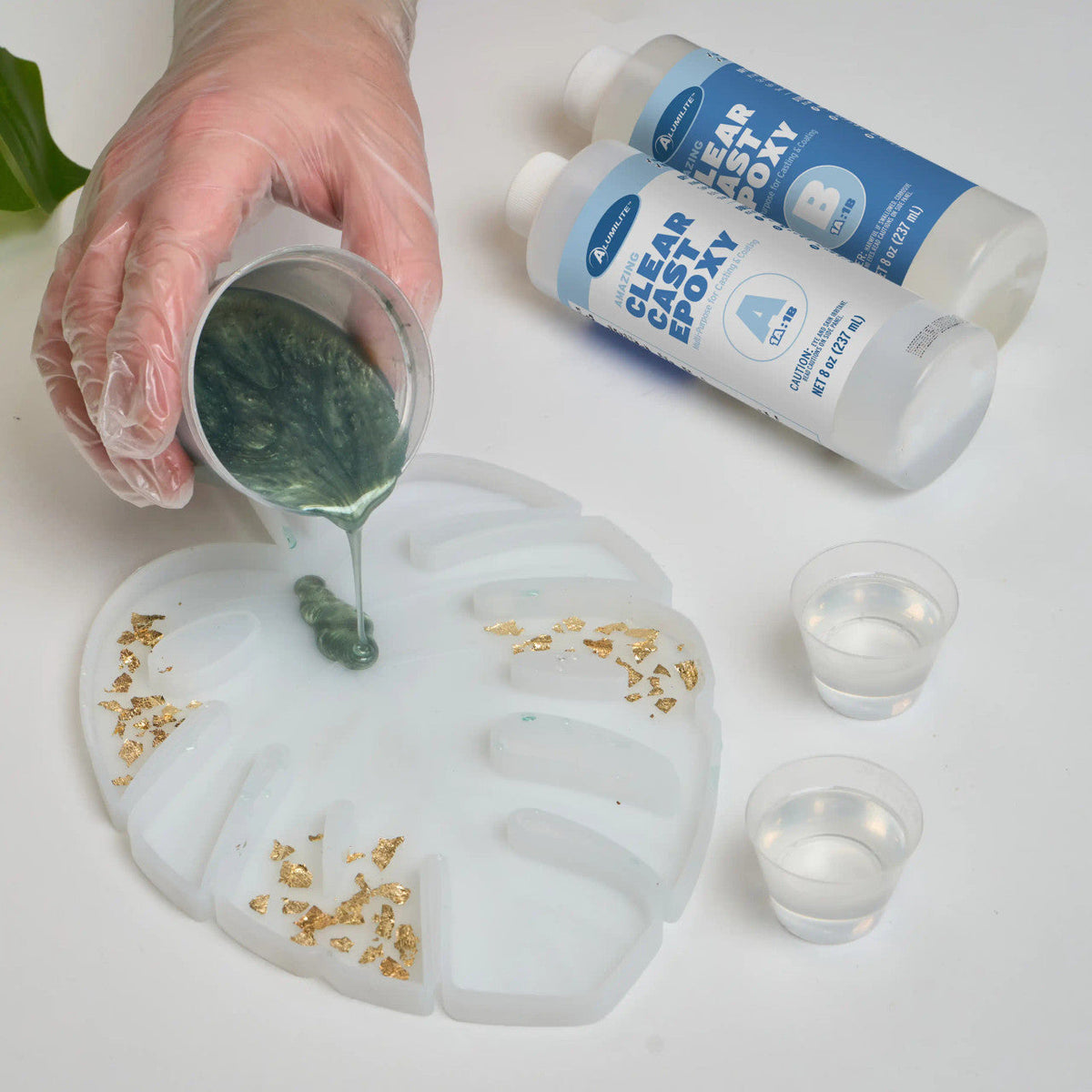 Gloved hand pours Notions’ Amazing Clear Cast Epoxy near gold flakes and plastic cups.