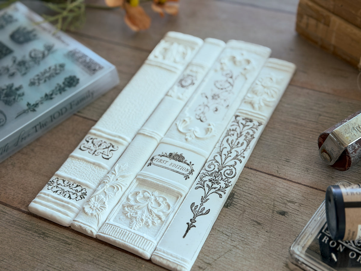 Four white, book spine-shaped I.O.D. The Connoisseur’s Library Mini Stamps arranged on a wooden surface.