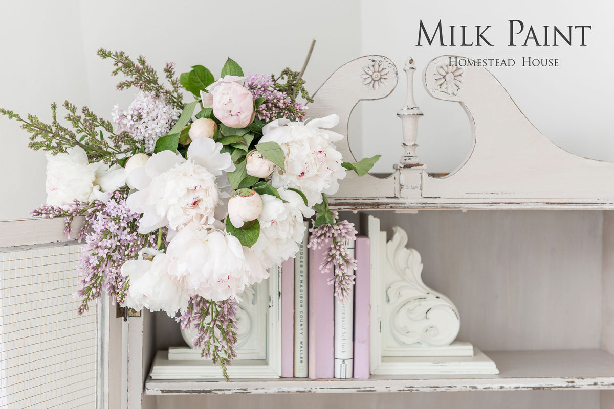 Homestead House Cathedral Taupe Milk Paint can with white and pink flowers and pastel books on a shelf.