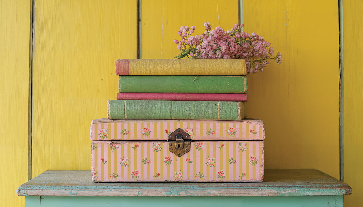 Box containing books and I.O.D. Ditsy Blooms Paint Inlay on a shelf, yellow paneled wall in background.