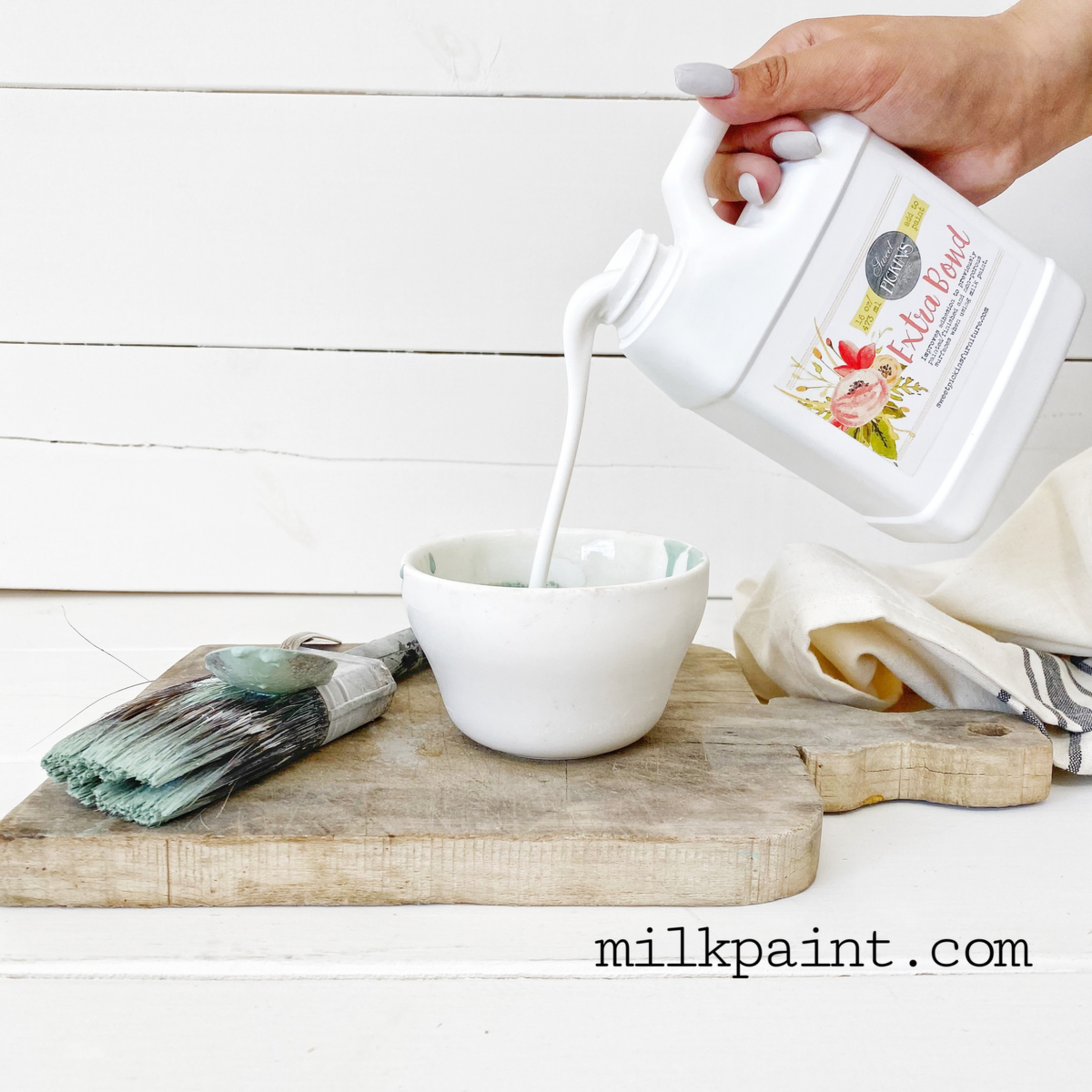 Hand pouring Sweet Pickins’ Extra Bond by Old Fashioned Milk Paint into a bowl, with paintbrush and cloth nearby.