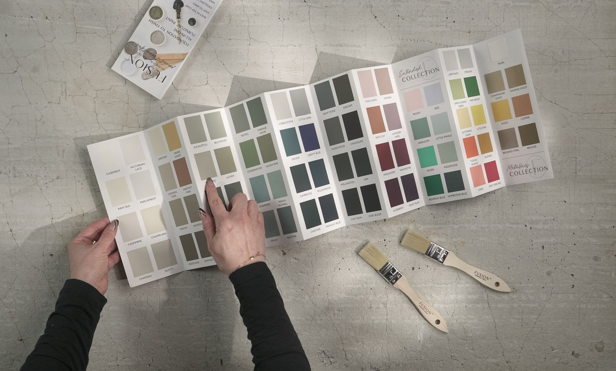 Hand pointing to Fusion Mineral Paint True to Color Card with real paint chips, brushes, and booklets nearby.