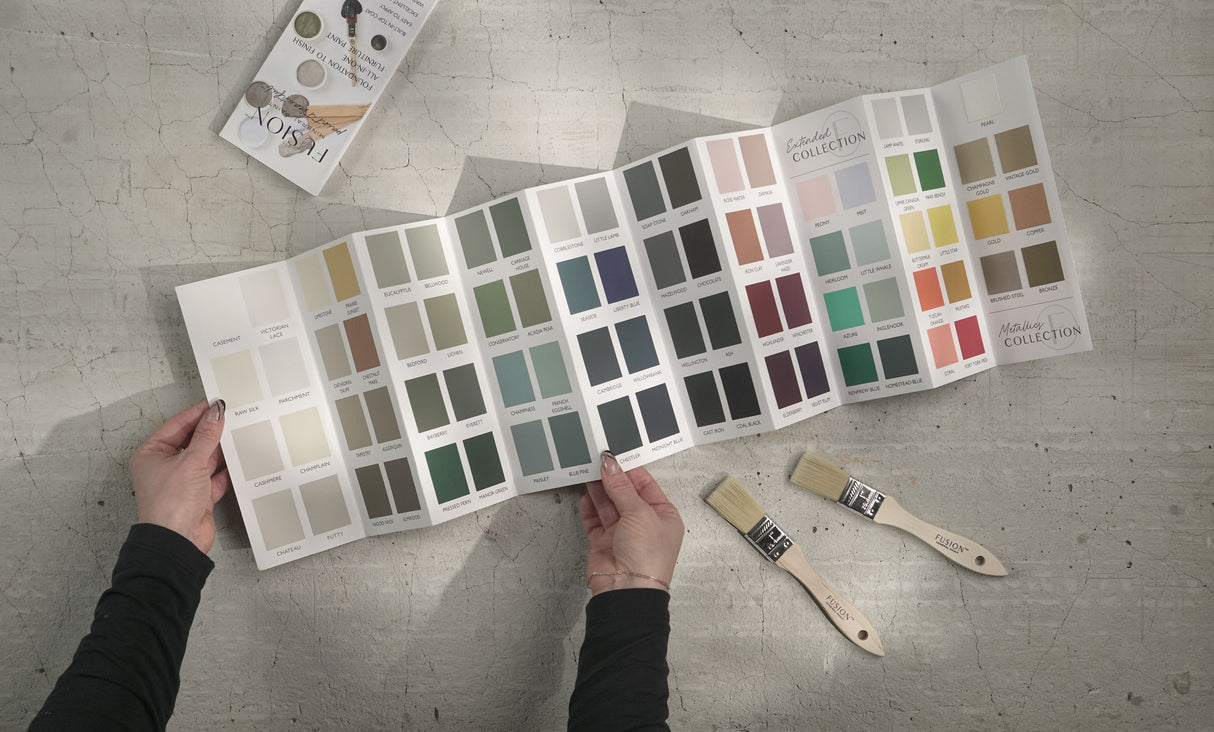 Hand holding Fusion Mineral Paint True to Color Card with 2024 paint chips, near paintbrushes and a magazine on gray.