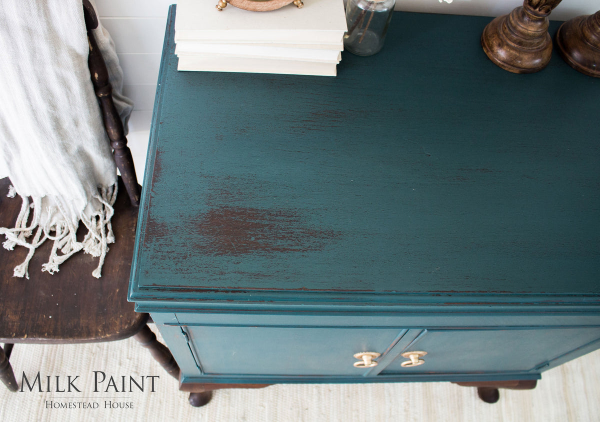 Teal wooden cabinet in Homestead House Gaspe Green Milk Paint displayed beside a dark chair and stack of books.