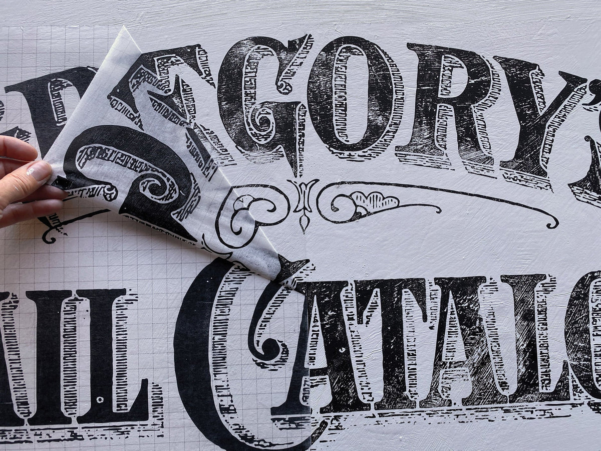 Hand lifting transfer paper to show Gregory’s Catalogue Paint Inlay by I.O.D. on a white background.