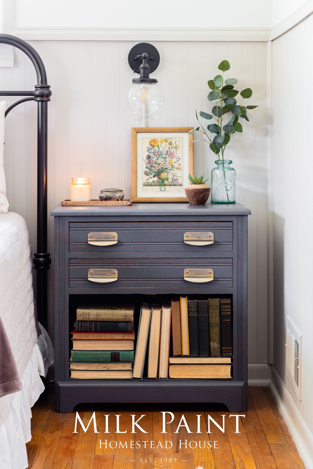 Homestead Grey Milk Paint nightstand with brass pulls, books, artwork, plant, candle, and light above.