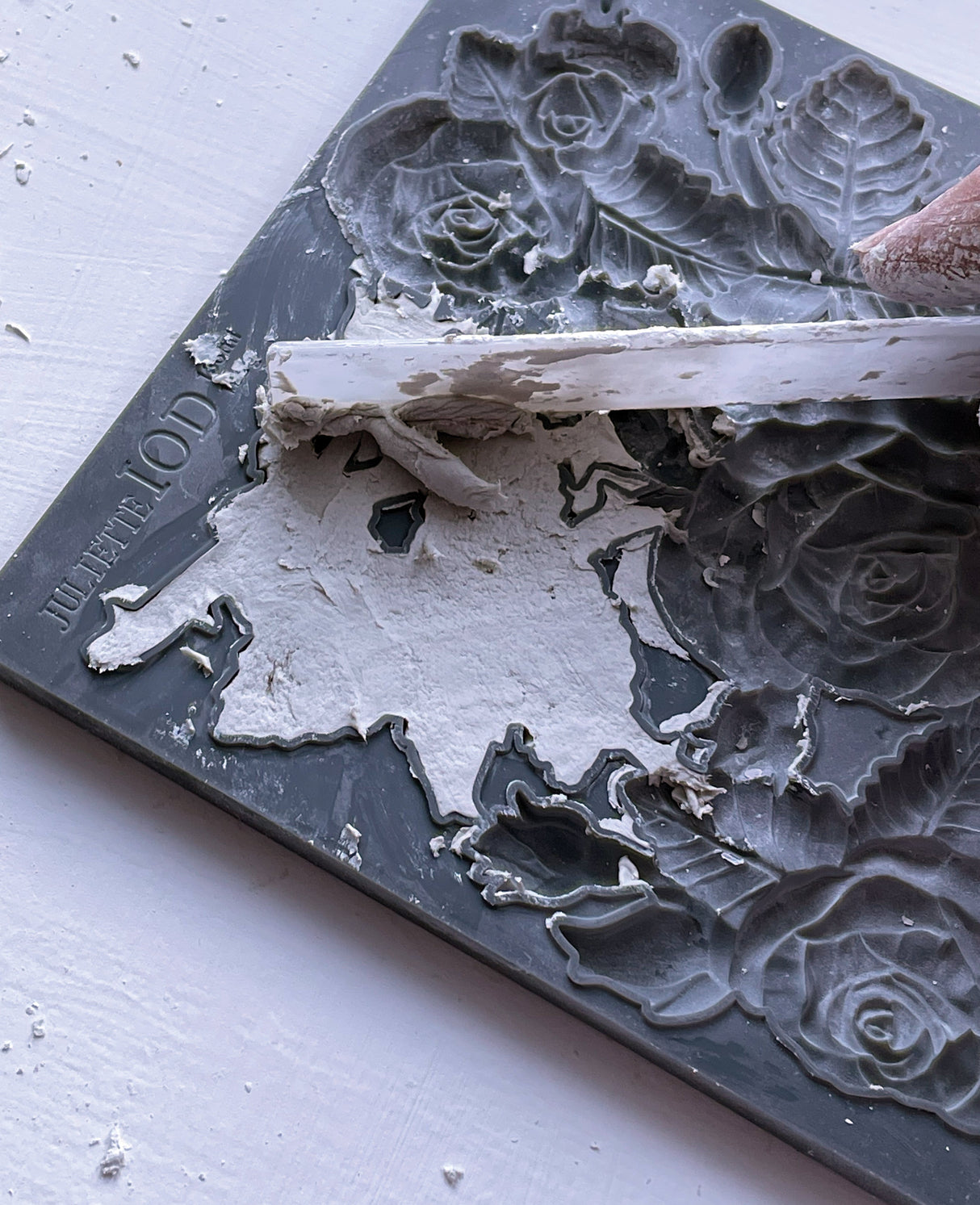 Hand scraping white plaster from I.O.D. Juliette Mould by Iron Orchid Designs on floral-patterned surface.
