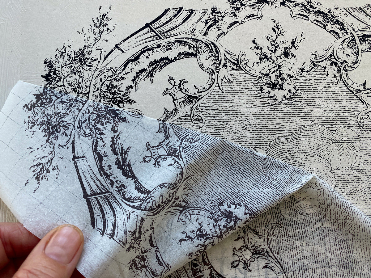 Hand holding fabric with black designs, resembling La Chasse Paint Inlay by IOD shown on decorated furniture below.