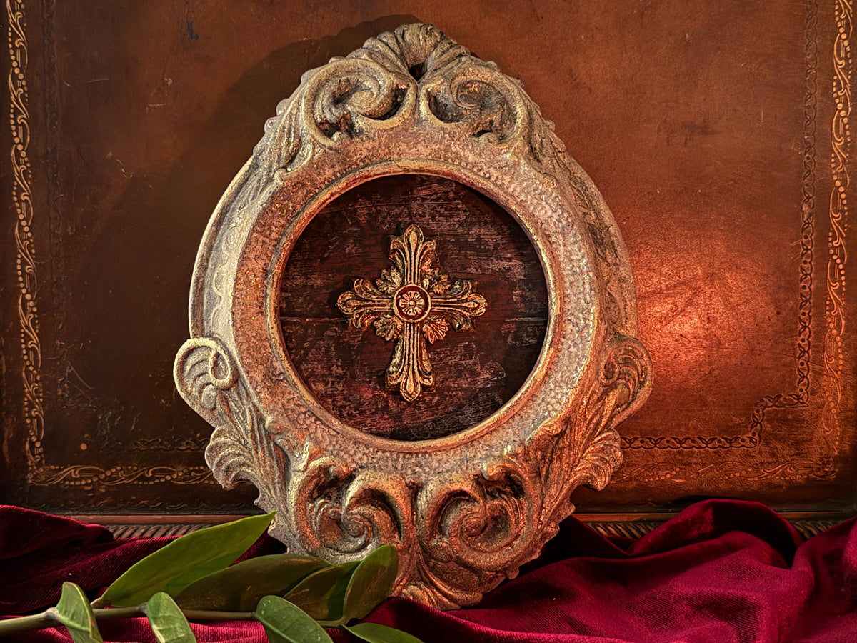 Ornate oval frame with gold I.O.D. Les Croix Mould cross on red fabric, green leaves in front.