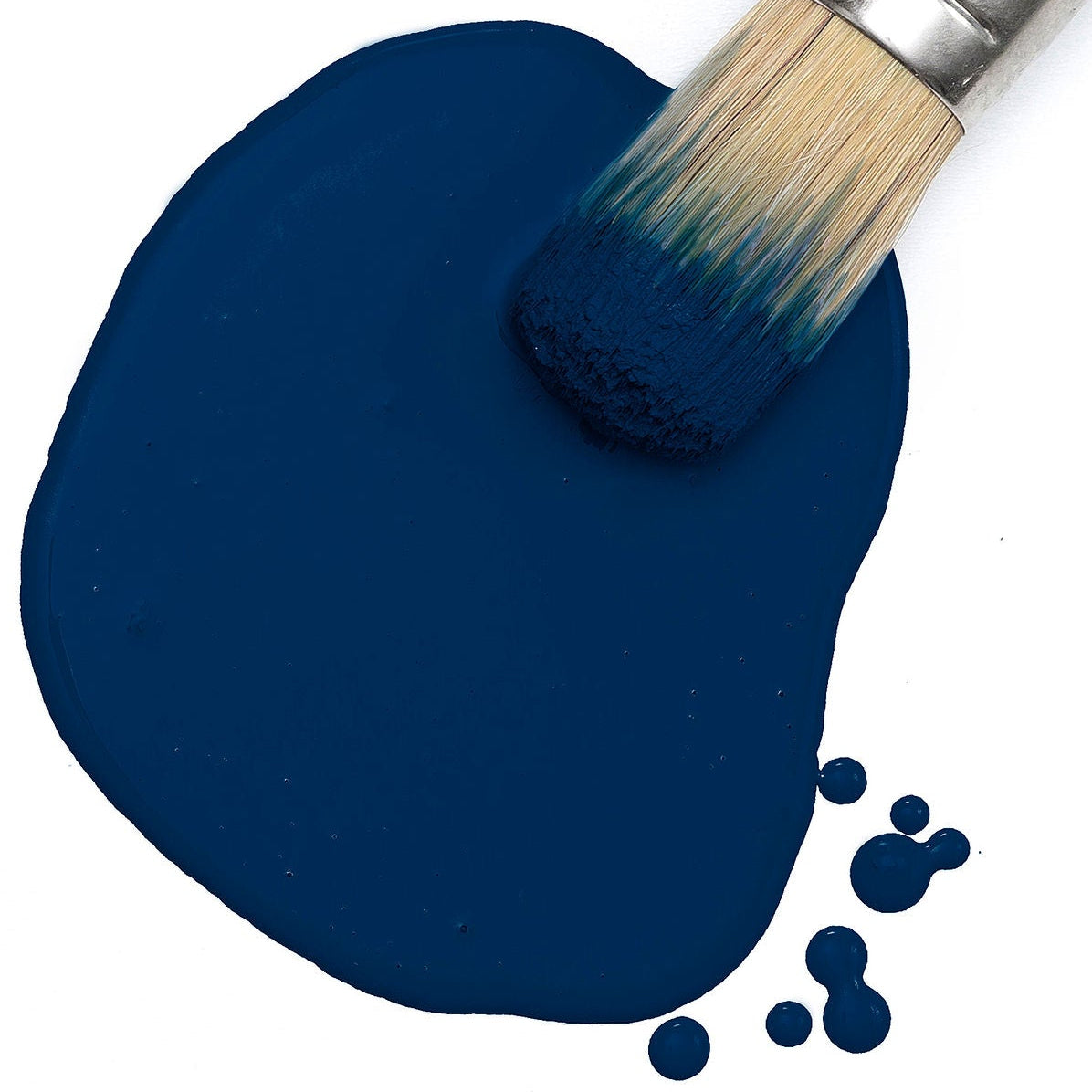 Paintbrush resting in Night Swim Milk Paint by Fusion Mineral Paint, dark blue on white surface with small drops nearby.