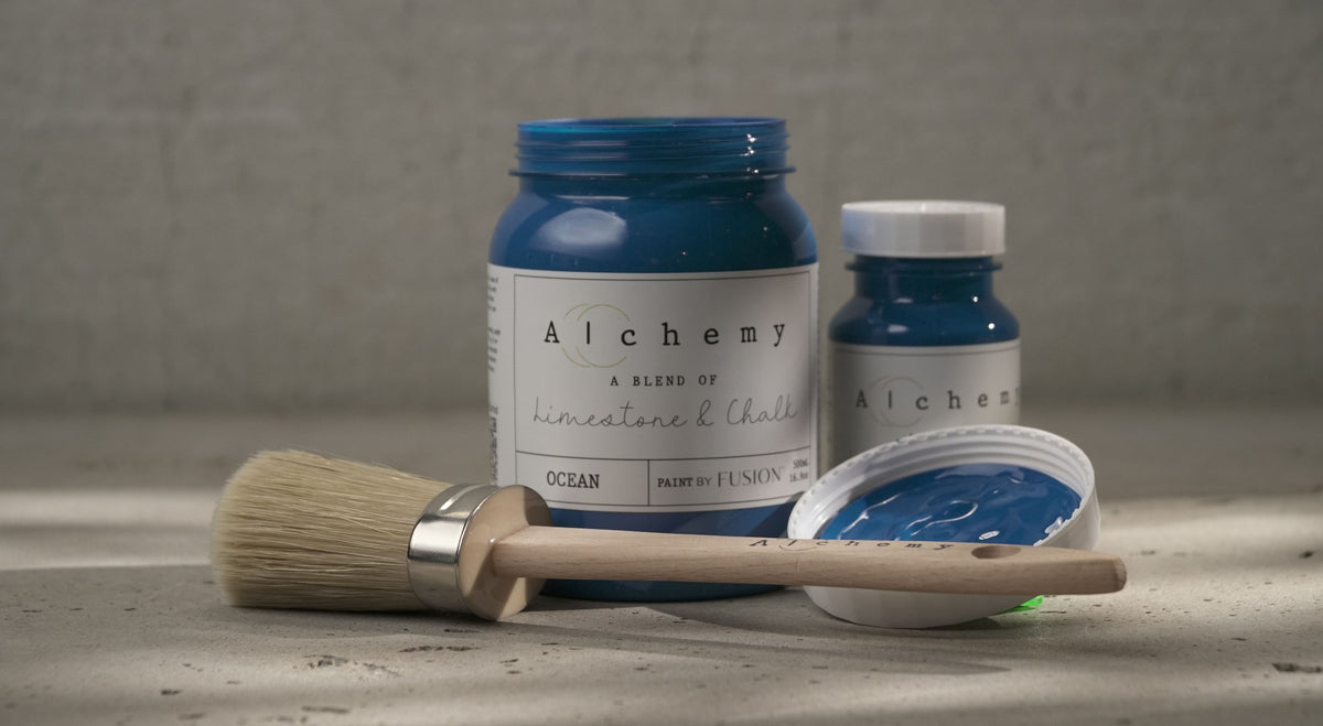 Paintbrush and two jars of Ocean Alchemy Paint by Fusion Mineral Paint on a textured surface in natural light.