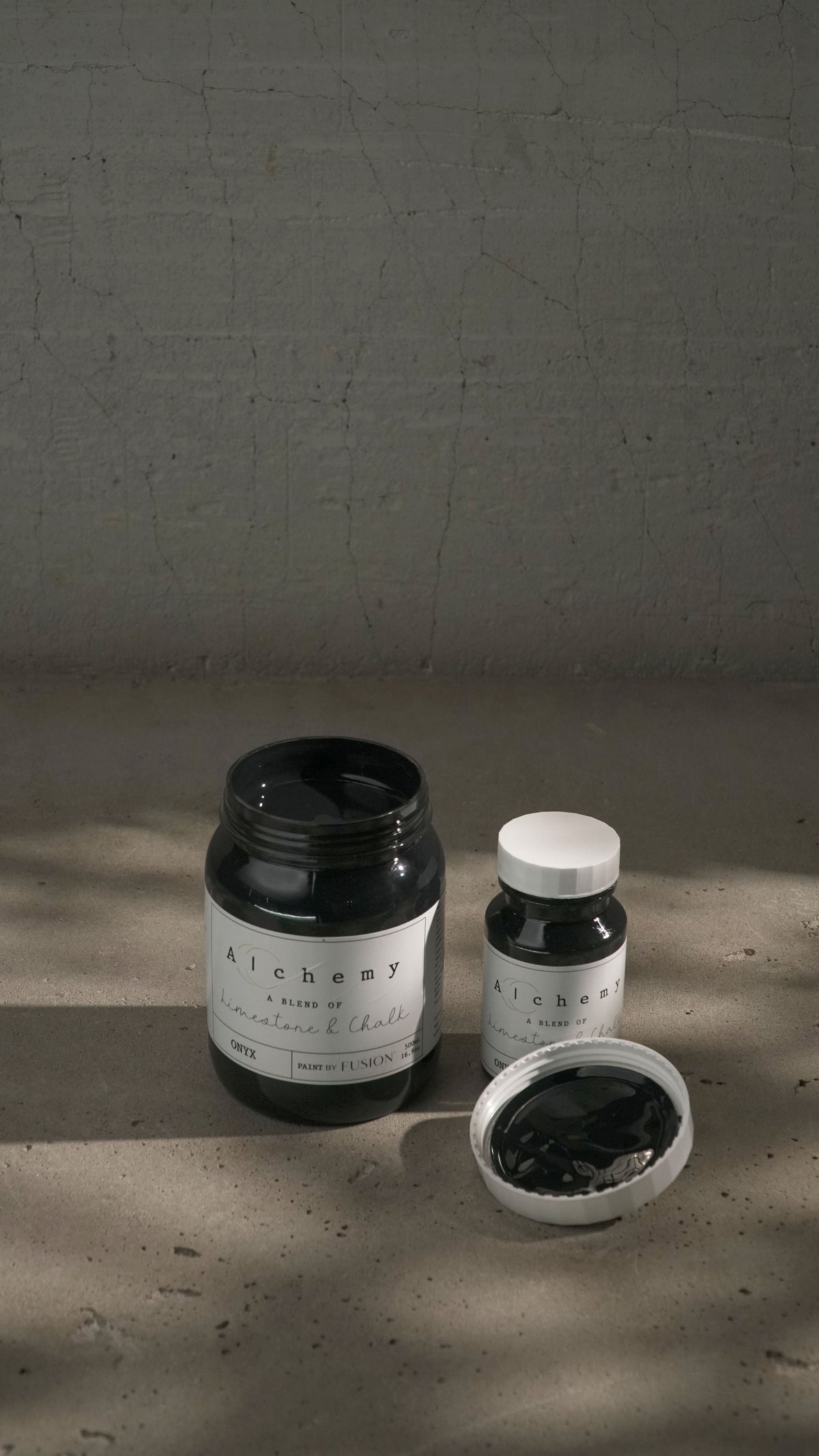 Two jars of Onyx Alchemy Paint by Fusion Mineral Paint displayed on a textured surface with dramatic lighting.