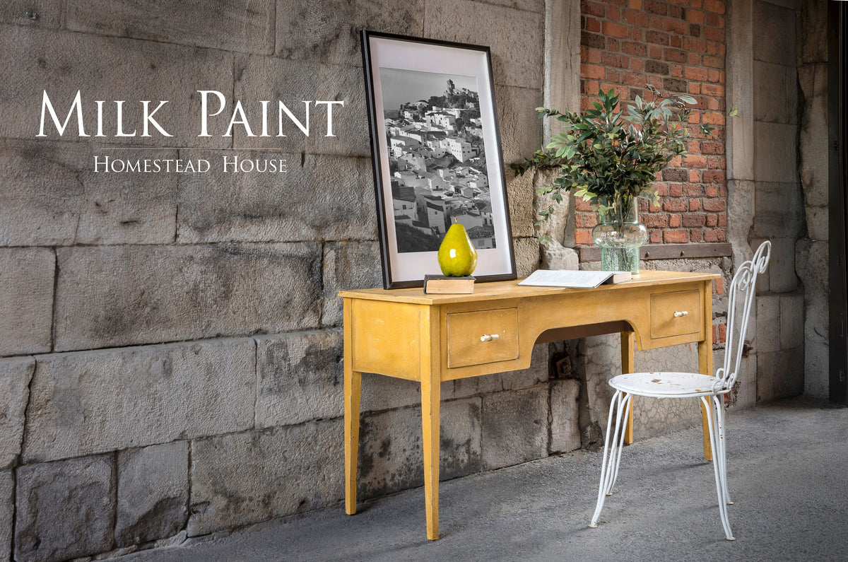 Yellow wooden desk in Prairie Sunset Milk Paint by Homestead House with green vase and plant near white chair.
