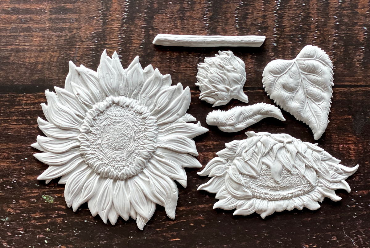 Six white clay I.O.D. Sunflowers Mould pieces and a leaf on a dark wood surface.