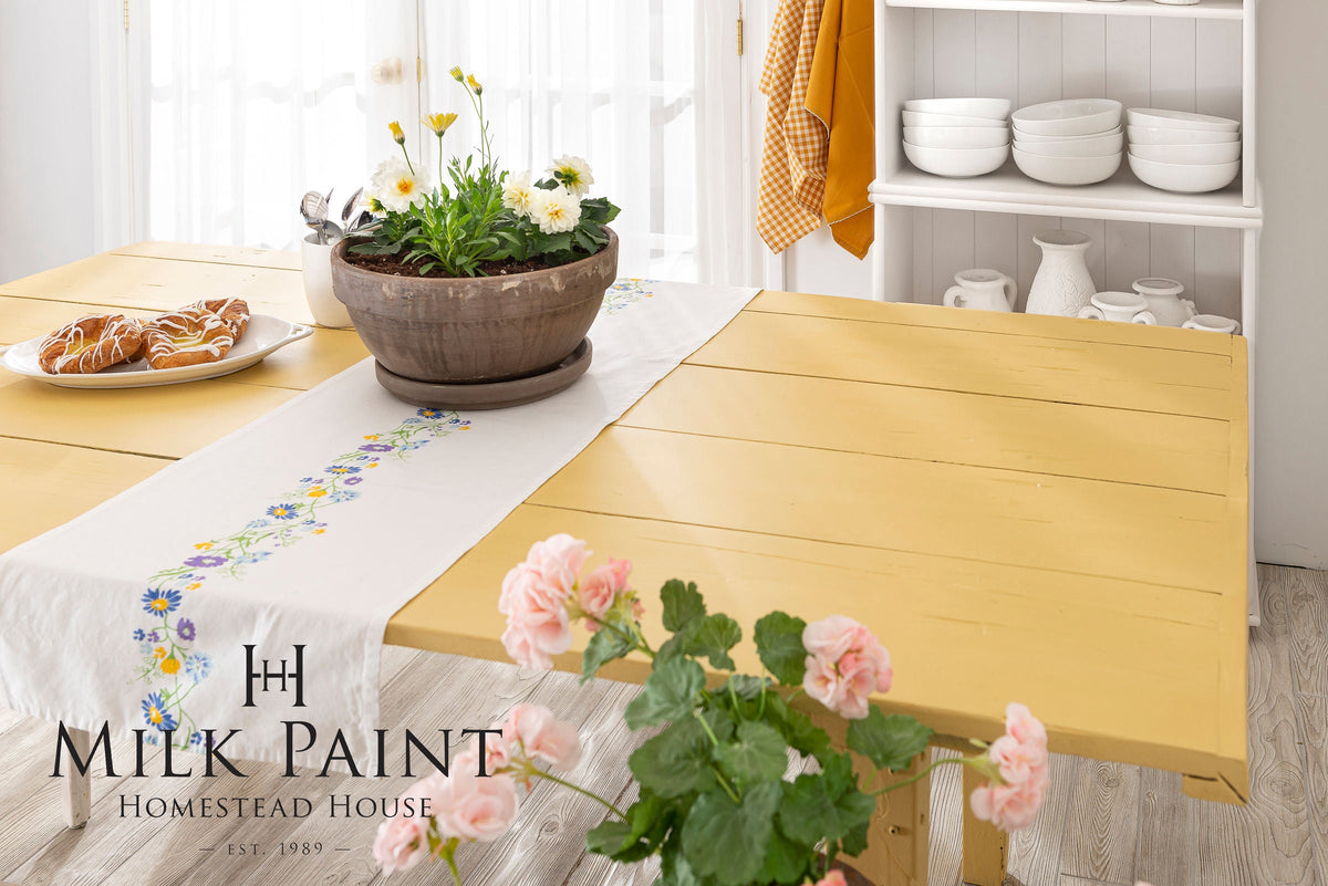 Yellow wood table painted with Homestead House Swedish Yellow Milk Paint, set with flowers, plates, and pastries.