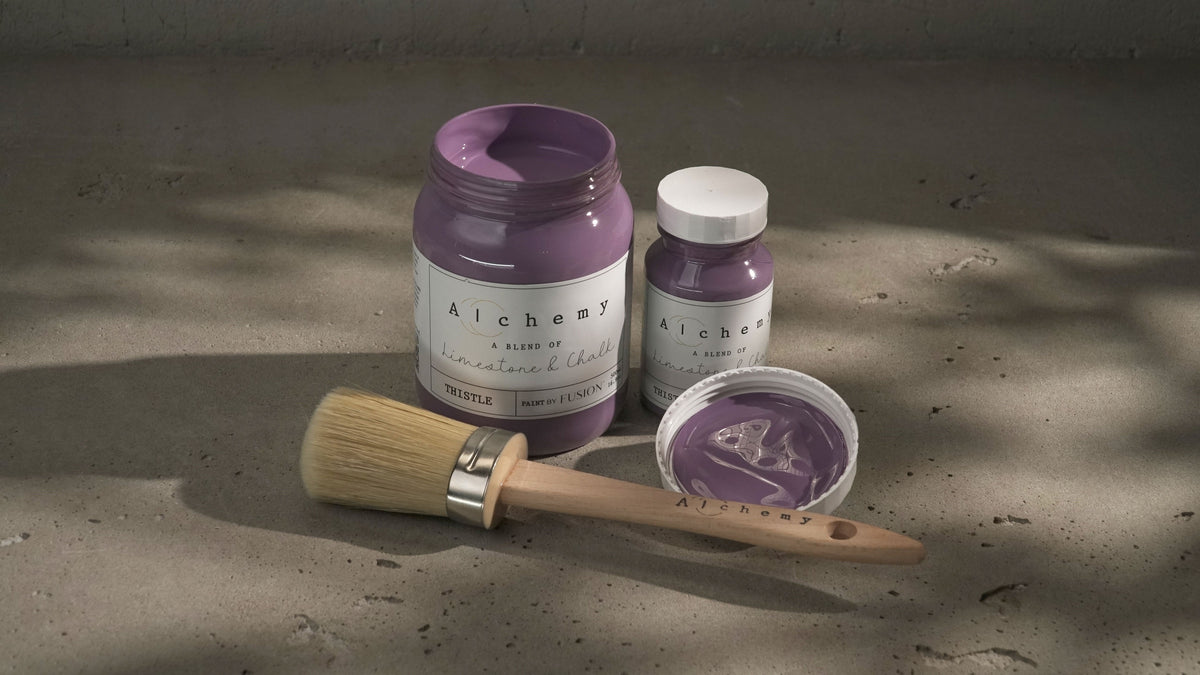 Two jars of Thistle Alchemy Paint by Fusion, one open with a brush resting nearby on a concrete surface.