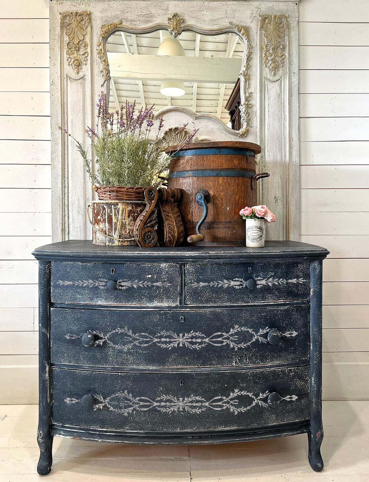 Distressed blue dresser with I.O.D. Trompe L’oeil Laurel Paint Inlay, topped with mirror, flowers, wood bucket and basket.