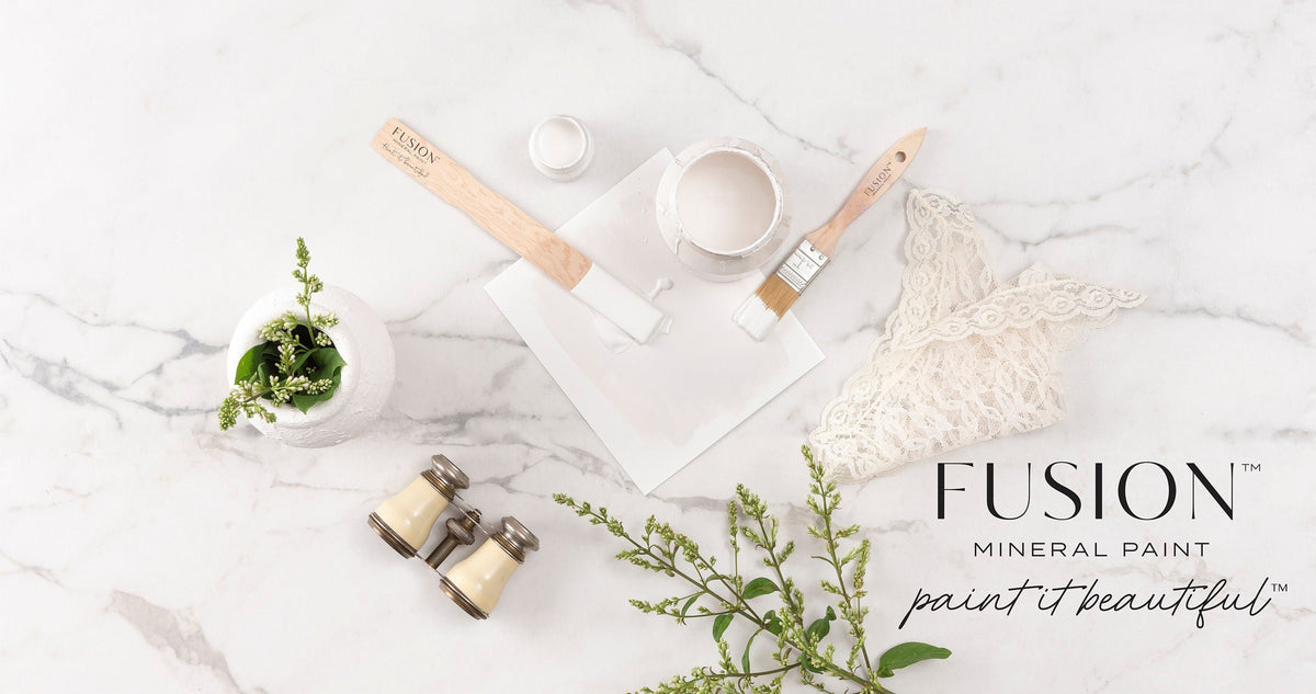 Fusion Mineral Paint Victorian Lace (multi-tone white) with paintbrushes, small plant, and binoculars on marble.