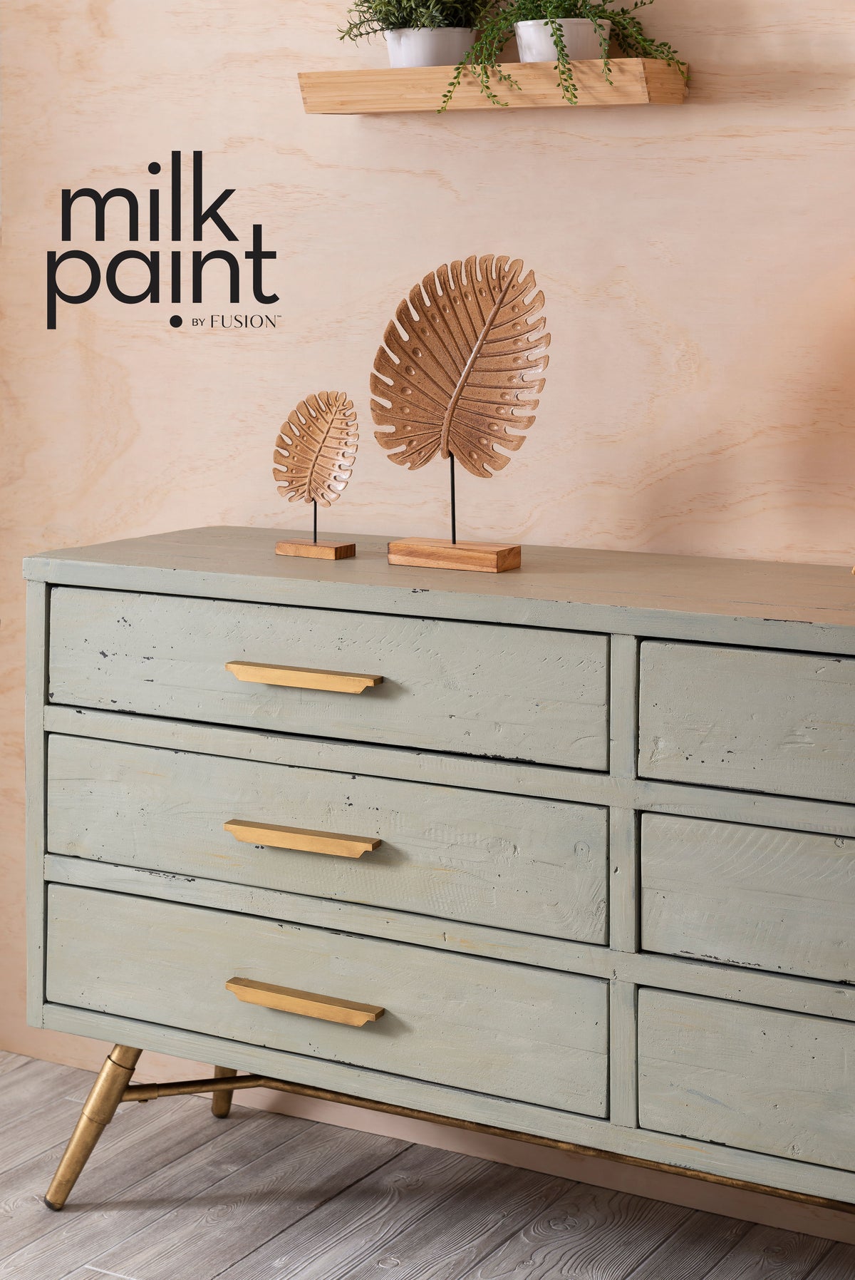 Sage green Fusion Mineral Paint Vintage Laurel dresser with gold handles, shown beneath leaf art and potted plants.