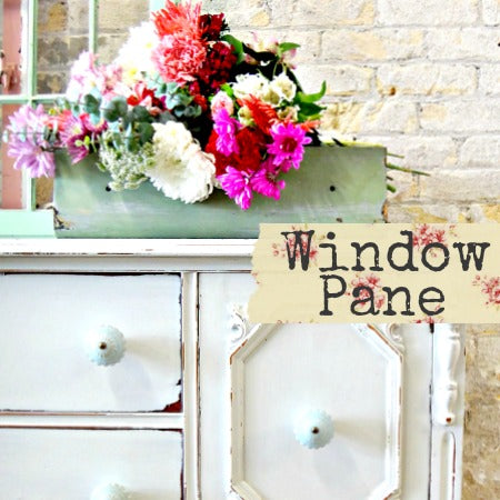 Green box of assorted flowers beside Old Fashioned Milk Paint Window Pane sign on white dresser.
