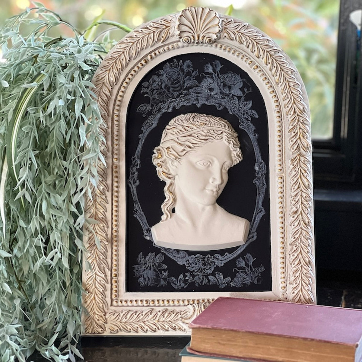Classical bust relief on I.O.D. Wood Gallery Blank with floral trim, next to green plant and books.