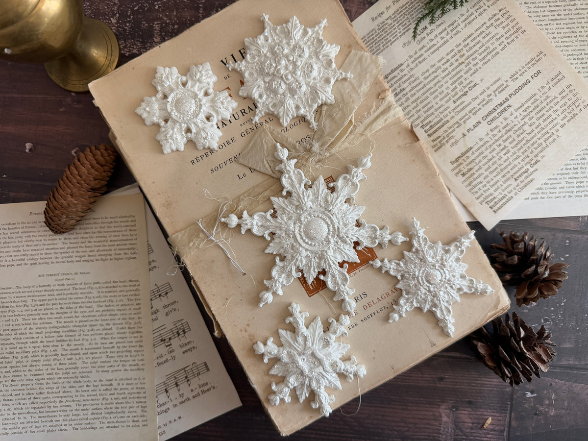 Four I.O.D. Snowflakes Mould (2026 Limited Release) ornaments displayed on a vintage book with pinecones nearby.