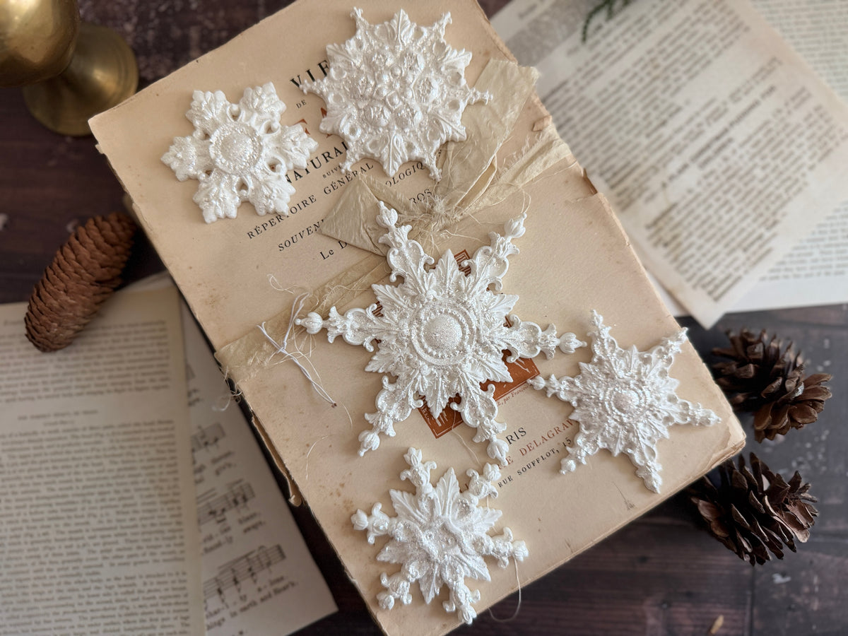 Five white snowflakes made with I.O.D. Snowflakes Mould (2026 Limited Release) on a vintage book background.