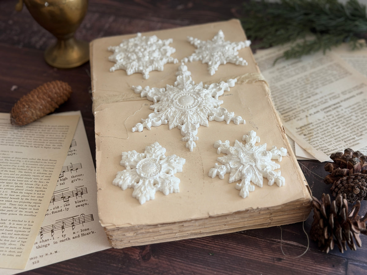 Five ornate snowflakes from the I.O.D. Snowflakes Mould (2026 Limited Release) displayed on an old book with pinecones.