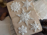 Five I.O.D. Snowflakes Mould (2026 Limited Release) ornaments displayed on a book with sheet music and pine cones.
