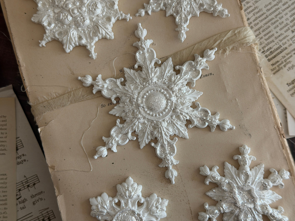 I.O.D. Snowflakes Mould (2026 Limited Release) white snowflake ornaments arranged on aged book paper with ribbon.