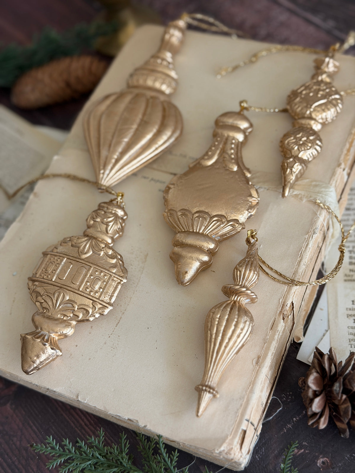 Five gold baubles made with I.O.D. Baubles Mould (2026 Limited Release) displayed on an old book with pinecones.