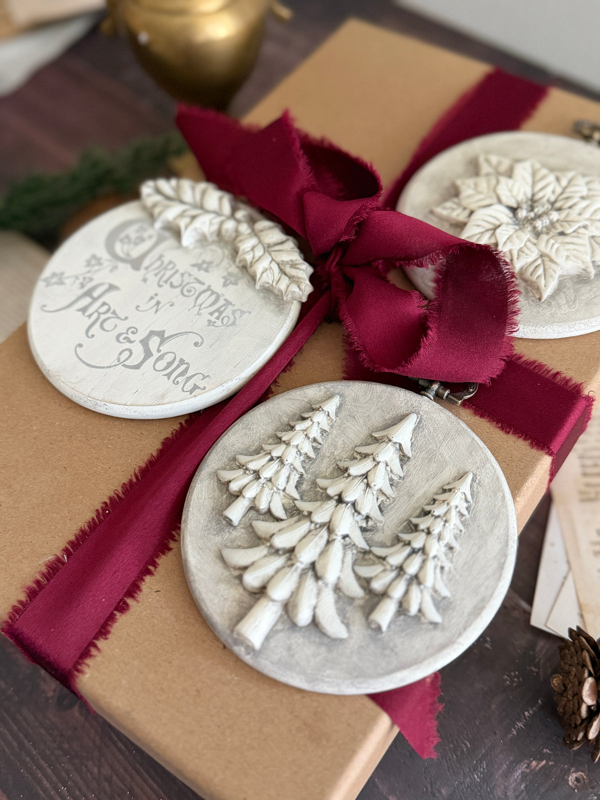 Brown gift box with dark red ribbon, topped with two I.O.D. Boughs of Holly ornaments (2026 Limited Release).