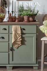 Green Acadia Pear Fusion Mineral Paint cabinet with checkered towel, copper accents, plants, and baskets on wood countertop.