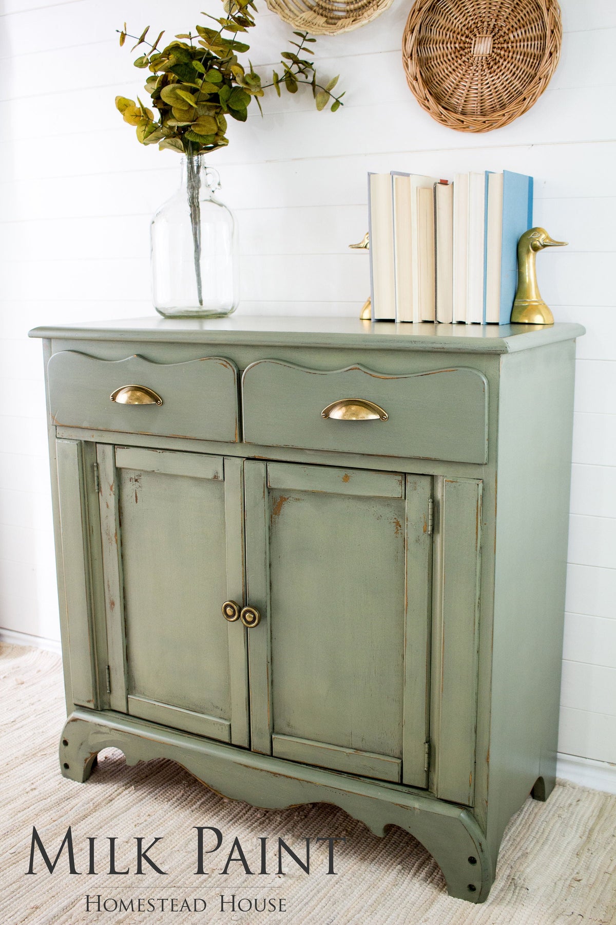 Homestead House Acadia Pear Milk Paint cabinet with brass pulls, topped with books, a vase, and duck bookends.