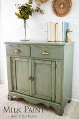 Homestead House Acadia Pear Milk Paint cabinet with brass pulls, topped with books, a vase, and duck bookends.