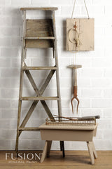 Rustic wooden ladder painted Algonquin by Fusion Mineral Paint beside beige stool against white brick wall.
