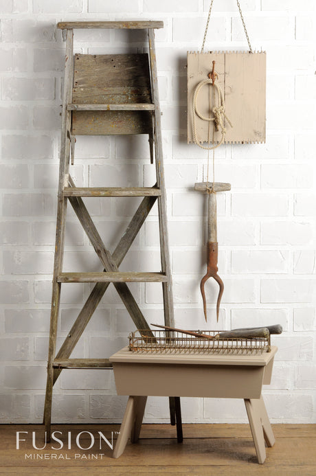 Rustic wooden ladder painted Algonquin by Fusion Mineral Paint beside beige stool against white brick wall.