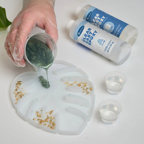 Gloved hand pours Notions’ Amazing Clear Cast Epoxy near gold flakes and plastic cups.