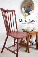 Chair painted Homestead House Barn Red Milk Paint next to stool holding cups, books, and a plant; sign above.
