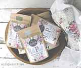Four bags of Basil Sweet Pickins Milk Paint by Old Fashioned Milk Paint with floral fabric and logo nearby.