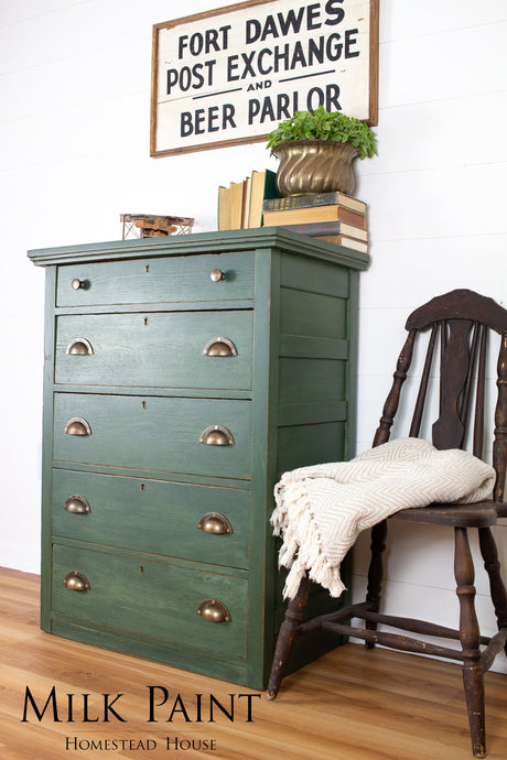 Green dresser in Homestead House Bayberry Milk Paint with brass handles, next to a wooden chair, books, and blanket.