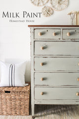 Light green dresser with round knobs painted with Homestead House Bedford Milk Paint, beside a woven basket and pillow.