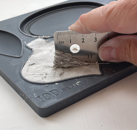 Hand smoothing white clay in Bibelots Mould (2024) by I.O.D. with metal ruler on gray tray.