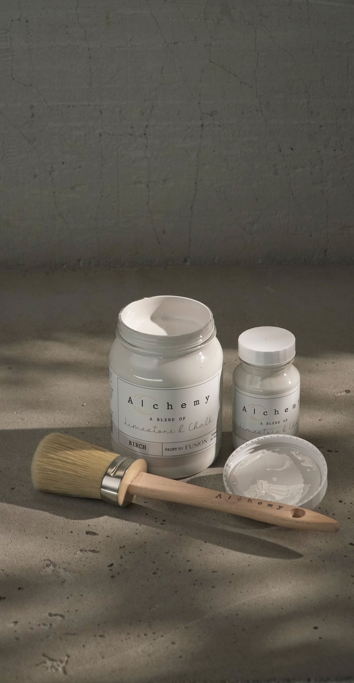 Two jars of Fusion Mineral Paint Birch Alchemy Paint, one open with a brush, showing labeled lids on a textured surface.