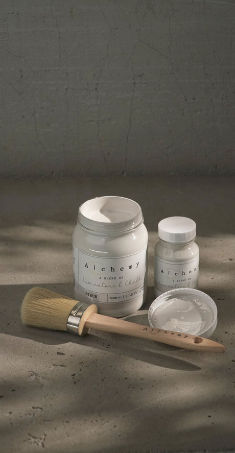 Two jars of Fusion Mineral Paint Birch Alchemy Paint, one open with a brush, showing labeled lids on a textured surface.