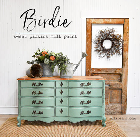 Green “Birdie” dresser by Old Fashioned Milk Paint, shown near flowers and a rustic door with a wreath.