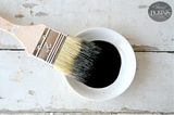 Paintbrush resting on a bowl of Sweet Pickins Black Oil Wax Old Fashioned Milk Paint on a white wooden surface.